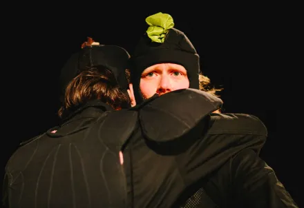 Zwei Schauspieler in schwarzen Kostümen umarmen sich auf der Bühne.
