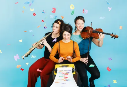 Drei Musikerinnen mit Schreibmaschine, Saxophon und Geige vor blauem Hintergrund.