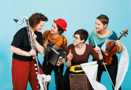 Vier Musikerinnen mit verschiedenen Instrumenten vor hellblauem Hintergrund.