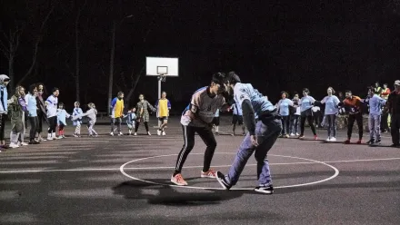 Zwei Personen stehen sich auf einem beleuchteten Basketballfeld gegenüber und beugen sich leicht nach vorne, als würden sie sich auf ein Spiel oder eine Aktion vorbereiten. Um sie herum bildet eine größere Gruppe einen Kreis, unter ihnen tanzende Kinder. Viele tragen helle Oberteile oder Westen. Die Szene spielt nachts, das Feld ist von Scheinwerfern erleuchtet.