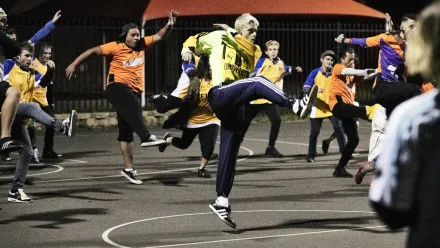 Mehrere Personen führen auf einem beleuchteten Außenplatz synchrone Sprünge mit angezogenen Knien aus. Die meisten tragen Sportkleidung in kräftigen Farben, darunter gelbe und orangefarbene Oberteile. Im Hintergrund sind ein Zaun und ein rotes Zeltdach zu sehen. Die Szene wirkt dynamisch, sportlich und tänzerisch.