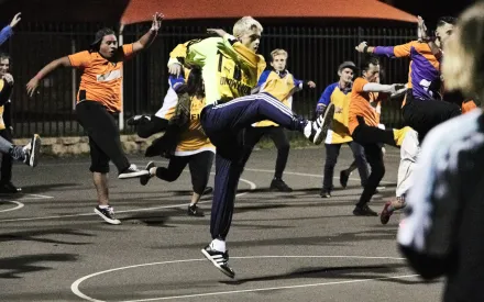 Mehrere Personen führen auf einem beleuchteten Außenplatz synchrone Sprünge mit angezogenen Knien aus. Die meisten tragen Sportkleidung in kräftigen Farben, darunter gelbe und orangefarbene Oberteile. Im Hintergrund sind ein Zaun und ein rotes Zeltdach zu sehen. Die Szene wirkt dynamisch, sportlich und tänzerisch.