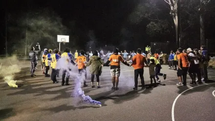 Menschen stehen nachts auf einem beleuchteten Basketballfeld im Kreis und halten sich an den Händen. Einige tragen bunte Trikots oder Jacken. Im Vordergrund steigt farbiger Rauch auf. Im Hintergrund ist ein Basketballkorb zu sehen, daneben stehen weitere Personen. Die Szene spielt sich im Freien ab, umgeben von Bäumen.