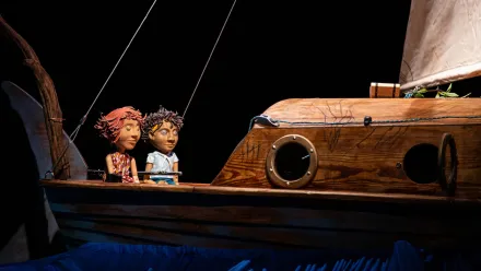 Zwei Puppen mit ausdrucksstarken Gesichtern sitzen nebeneinander auf dem Deck eines detailreich gestalteten Holzboots. Die Puppen tragen bunte Kleidung und blicken nach vorne. Das Boot besitzt runde Bullaugen und ein großes, weißes Segel. Im Vordergrund sind stilisierte blaue Wellen zu erkennen, der Hintergrund ist dunkel gehalten.