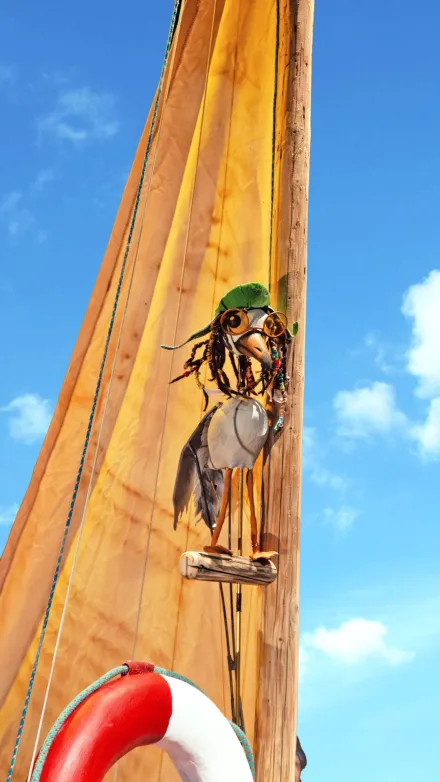 Im Vordergrund ist eine hölzerne Skulptur in Form eines Vogels zu sehen, die auf einem Segelmast sitzt. Die Skulptur besteht aus verschiedenen Materialien wie Holz, Metall und Seil, und trägt eine grüne Kopfbedeckung. Im Hintergrund ragt ein gelbes Segel schräg nach oben, darüber ein blauer Himmel mit wenigen weißen Wolken. Am unteren Bildrand ist ein Teil eines Rettungsrings mit rotem und weißem Muster sichtbar.
