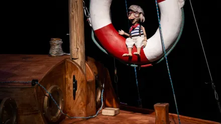 Im Mittelpunkt hängt eine Gliederpuppe mit Brille, gestreiftem Oberteil und kurzen Hosen in einem großen Rettungsring mit rotem und weißem Muster. Die Szene spielt auf einem hölzernen Schiffsdeck mit sichtbaren Bullaugen, Seilen und einem kleinen Holzkasten. Der Hintergrund ist dunkel gehalten, sodass die Figuren und das Deck deutlich hervortreten.