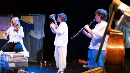 Vier Personen musizieren auf einer Bühne. Links kniet eine Person auf einem Stapel bunter Textilien und spielt Geige. Daneben steht eine Person in weißer Kleidung und spielt ein Blechblasinstrument. Rechts davon spielt eine weitere Person Klarinette. Am rechten Bildrand ist ein Kontrabass und ein Teil einer weiteren Person zu sehen. Im Hintergrund hängt eine Konstruktion aus Stoffbahnen und Kleidungsstücken. Die Bühne ist dunkel, die Szene wird von Scheinwerfern beleuchtet.