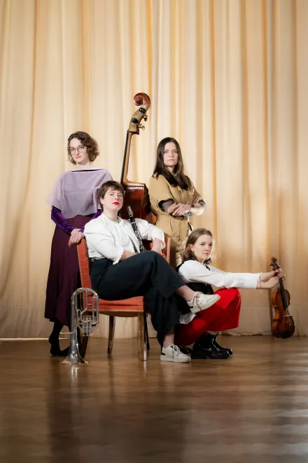 Vier Personen mit klassischen Musikinstrumenten: Kontrabass, Posaune und Geige, vor einem beigen Vorhang in einem Raum mit Holzboden