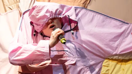 Mittig zu sehen ist eine Person hinter einem Vorhang aus bunten Textilien. Durch eine rosa Kapuzenjacke hat die Person den Kopf und einen Arm gesteckt, die Hand hält ein Mikrofon zum Mund, die Kapuze der Jacke hängt bis zur Nase über das Gesicht, sodass man nur Mund und Nase sieht