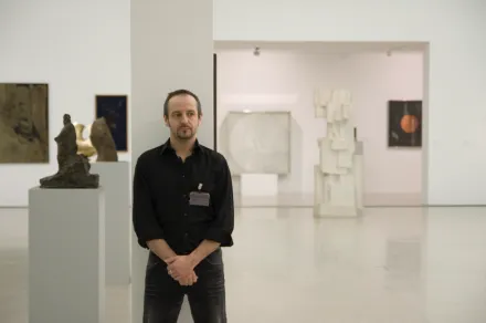 Mann in schwarzem Hemd und Jeans steht mit verschränkten Händen in einer Galerie mit moderner Kunst und Skulpturen