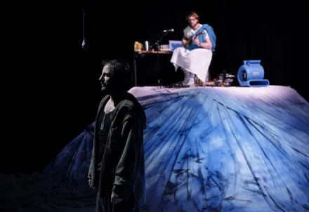 Two actors on stage with a blue fabric landscape; one stands in front, the other plays guitar in the background.