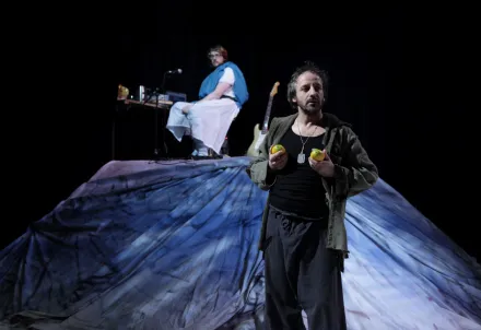 Schauspieler steht mit Zitronen in den Händen vor einer blauen Bühnenlandschaft, Musiker sitzt dahinter.