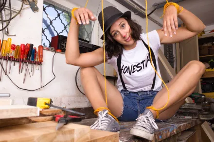 Person sitzt auf Werkbank mit hochgezogenen Beinen, trägt weißen T-Shirt mit Aufschrift 'HONESTY', Jeansshorts, Hosenträger und schwarzen Hut, hält gelbe Marionettenfäden