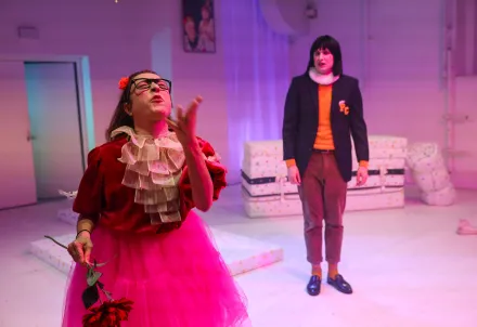 in pinkem Tüll gekleidete Prinzessin mit schwarzer Hornbrille, die wild gestikulierend Richtung Publikum spricht, im Hintergrund ein verloren wirkender Prinz mit Sakko und Mühlradkragen, die Prinzessin ansehend