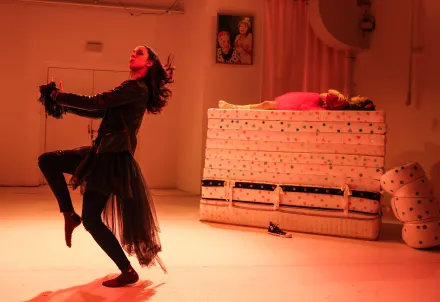 eine prinzessin schlafend auf einem matratzenstapel im hintergrund, eine schwarz gekleidete junge frau im vordergrund beim hobby horsing