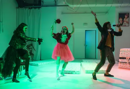 drei personen, wild tanzend und singend, im Hintergrund ein Stapel Matratzen und ein Bild mit einem König und einer Königin