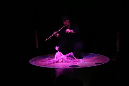 Eine Marionette sitzt auf einem beleuchteten runden Podest, eine Person steuert sie mit Stäben im Hintergrund