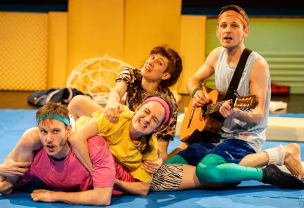 Vier Darsteller:innen in bunten Outfits performen energiegeladen auf blauer Matte.