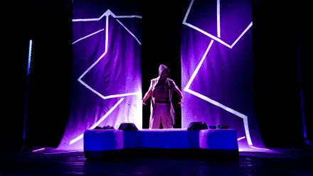 Eine Person steht mittig auf einer Bühne, trägt offen eine ärmellose Kapuzenweste und helle Kleidung. Im Hintergrund hängen zwei große Stoffbahnen mit leuchtenden, geometrischen Linien in Violett- und Blautönen. Die Bühne ist in dunkles Licht getaucht, das von violetten und blauen Akzenten durchbrochen wird. Im Vordergrund befindet sich ein rechteckiges Podest.