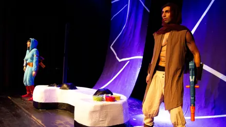 Zwei Personen stehen auf einer Theaterbühne mit abstrakten, geschwungenen Kulissenelementen in dunklem Blau und Weiß. Die Person im Vordergrund trägt eine ärmellose braune Weste, eine beigefarbene Hose, einen weinroten Schal und hält ein großes, blaues Requisit in der Hand. Die Person im Hintergrund trägt ein hellblaues Kostüm mit Kapuze und roten Stiefeln. In der Bühnenmitte liegt ein überdimensionaler Controller. Die Szene ist farbenfroh ausgeleuchtet.