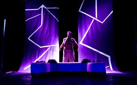 Eine Person steht mittig auf einer Bühne, trägt offen eine ärmellose Kapuzenweste und helle Kleidung. Im Hintergrund hängen zwei große Stoffbahnen mit leuchtenden, geometrischen Linien in Violett- und Blautönen. Die Bühne ist in dunkles Licht getaucht, das von violetten und blauen Akzenten durchbrochen wird. Im Vordergrund befindet sich ein rechteckiges Podest.