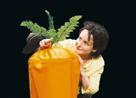 Person in gelbem Hemd hält eine orangefarbene Box mit grünen Farnblättern und einer Schneckenfigur darauf