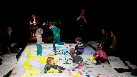 Mehrere Kinder spielen auf einer großen, weißen Bodenfläche, die mit bunten Bausteinen und gelben Stoffstücken bedeckt ist. Einige Kinder sitzen, andere bewegen sich laufend oder krabbelnd. Im Hintergrund sitzen und stehen Erwachsene am Rand der Spielfläche, eine Person spielt ein Blasinstrument. Die Szene ist in einen dunklen Raum getaucht, das Licht konzentriert sich auf die Spielfläche.