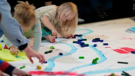 Ein Kind mit blondem Haar kniet auf einem großflächigen Papierbogen, der mit blauen Linien und roten Flächen bemalt ist. Es greift nach kleinen, rechteckigen Bausteinen, die in verschiedenen Farben auf dem Papier verteilt liegen. Weitere Kinderhände und -arme sind am Rand sichtbar, ebenfalls mit Bausteinen beschäftigt. Die Szene wirkt wie ein gemeinsames kreatives Spiel oder eine Bastelaktion auf dem Boden.
