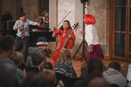 Kinderkonzert mit Musikerin am Cello und zwei Darstellern, eine trägt rote Perücke und karierte Rock, Publikum aus Kindern sitzt im Vordergrund