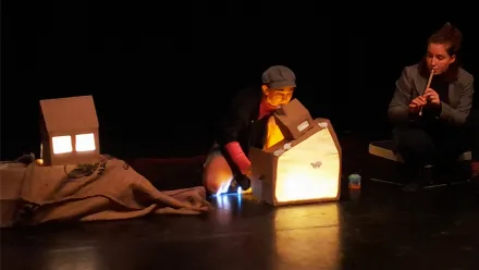Eine dunkle Bühne, auf der kleine von innen leuchtende Häuschen zu sehen sind, am rechten Bildrand sitzt eine Person und spielt Flöte, daneben kniet eine Person mit Wollkappe, macht ein Schattenspiel an der Wand eines der beleuchteten Häuschen.