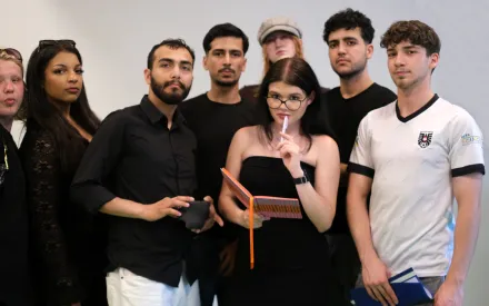 Gruppenfoto mit acht jungen Menschen, bis auf einen tragen alle schwarze Oberteile. Eine Person hat ein Notizbuch und einen Bleistift in der Hand, den Bleistift hält sie an die Lippen