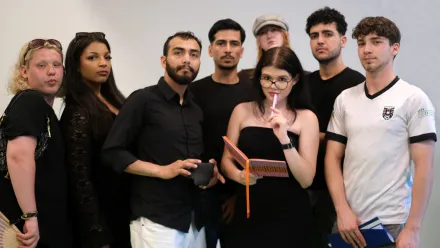 Gruppenfoto mit acht jungen Menschen, bis auf einen tragen alle schwarze Oberteile. Eine Person hat ein Notizbuch und einen Bleistift in der Hand, den Bleistift hält sie an die Lippen