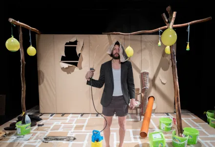 Performer auf der Bühne in einer surrealen Szenerie mit Eimern, Ballons und Papierwänden.