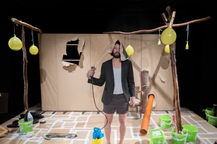 Person steht auf einer Bühne mit schwarzem Blazer, grauem T-Shirt und Shorts, hält eine Angelrute, umgeben von gelben Luftballons und grünen Eimern