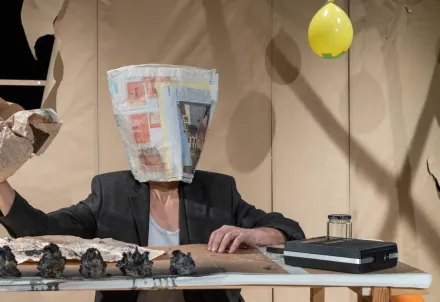 Eine Person mit Papiertüte über dem Kopf sitzt an einem Tisch voller Papier und Ballons.