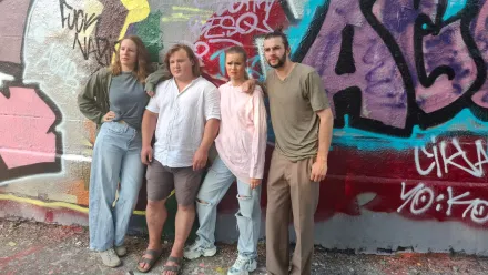 Vier Personen stehen nebeneinander vor einer Wand mit bunter Graffitibemalung. Die linke Person trägt eine helle Jacke und eine weite Jeans, die zweite Person ein weißes Hemd und kurze Hosen, die dritte Person ein rosa Sweatshirt und eine zerrissene Jeans, die rechte Person ein beigefarbenes T-Shirt und eine dunkle Hose. Alle blicken in Richtung Kamera, die Körperhaltung wirkt entspannt. Der Boden besteht aus Kies oder Schotter.