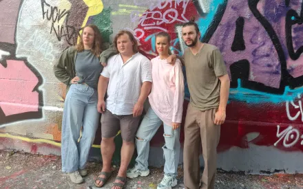 Vier Personen stehen nebeneinander vor einer Wand mit bunter Graffitibemalung. Die linke Person trägt eine helle Jacke und eine weite Jeans, die zweite Person ein weißes Hemd und kurze Hosen, die dritte Person ein rosa Sweatshirt und eine zerrissene Jeans, die rechte Person ein beigefarbenes T-Shirt und eine dunkle Hose. Alle blicken in Richtung Kamera, die Körperhaltung wirkt entspannt. Der Boden besteht aus Kies oder Schotter.