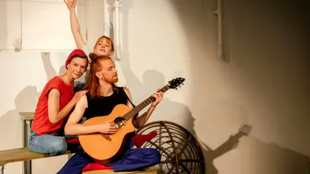 Drei Personen sitzen auf unterschiedlich hohen Flächen beeinander, die Person ganz vorne spielt Gitarre. Der Raum, in dem sie sich befinden, ist weiß, ihre Kleidung ist farbig rot, blau, schwarz.
