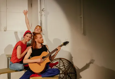 Drei Performer*innen in einer ruhigen, harmonischen Szene mit Gitarre und sanftem Licht.