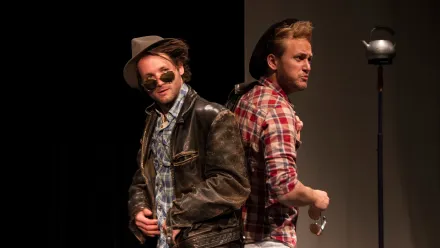 zwei junge männer stehen mit den Rücken aneinandergelehnt in der Mitte des Fotos. Beide tragen einen Hut, der eine ein rot-beige kariertes Hemd, der andere eine Lederjacke und Sonnenbrille. Der in der Lederjacke gibt sich "cool und abgebrüht", der andere fragend.