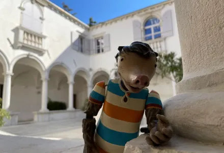 Eine Maulwurf-Figur in gestreiftem Shirt lehnt an einer Säule in einem sonnigen Innenhof.