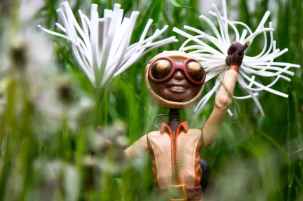 Skulptur einer lächelnden Figur mit großen runden Brillen und orangefarbenem Anzug, die eine weiße Blume hält