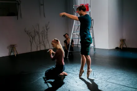 Tänzerin kniet auf dem Boden und spielt Flöte, Tänzer steht auf Zehenspitzen mit erhobenen Armen in einem dunklen Tanzstudio