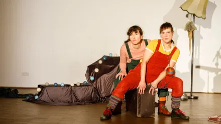 Zwei Personen sitzen auf einem dunklen Kasten in einem hell ausgeleuchteten Raum. Beide tragen bunte Latzhosen, gestreifte Socken und dunkle Schuhe. Die linke Person trägt eine rosa Oberteil, die rechte ein gelbes. Im Hintergrund liegt ein mit Stoff bedeckter Gegenstand, auf dem zahlreiche runde, helle und blaue Kugeln verteilt sind. Rechts steht eine Stehlampe mit hellem Lampenschirm. Der Boden ist aus Holz, die Wand im Hintergrund ist weiß.