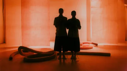 Zwei Personen stehen nebeneinander in einem Raum mit orangefarbener Beleuchtung. Sie sind von hinten zu sehen und werfen lange Schatten auf den Boden. Im Hintergrund befinden sich große, helle Flächen und mehrere dicke, schlangenförmige Objekte, die auf dem Boden liegen. Die Szene wirkt minimalistisch und ruhig.