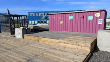 Freiluft-Bühne mit Holzboden, im Bühnenhintergrund ein altrosa gestrichener Baucontainer mit bunten Sprechblasen darauf